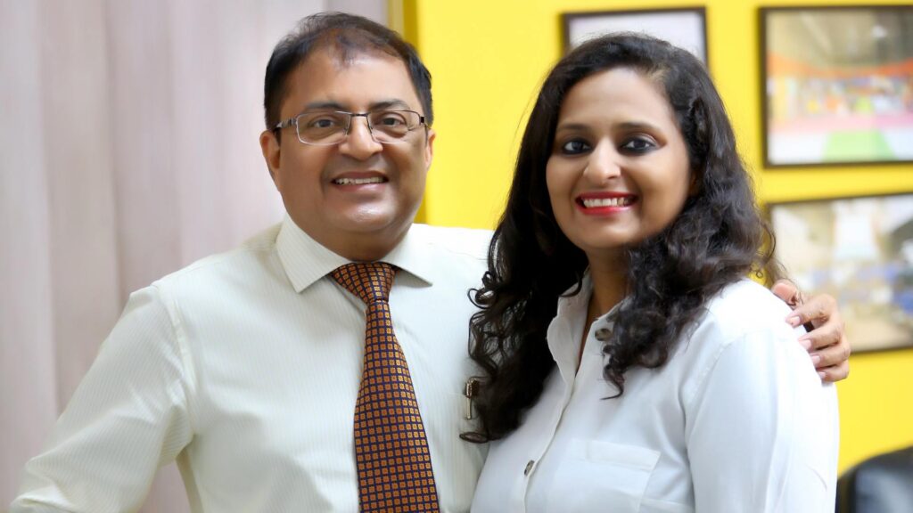 Aakanksha Bhargava stands beside her father, Rajeev Bhargava, both smiling confidently in a warmly lit office with framed memories on the yellow wall behind them. Their professional yet affectionate posture captures the essence of balancing tradition and innovation while leading a family legacy.