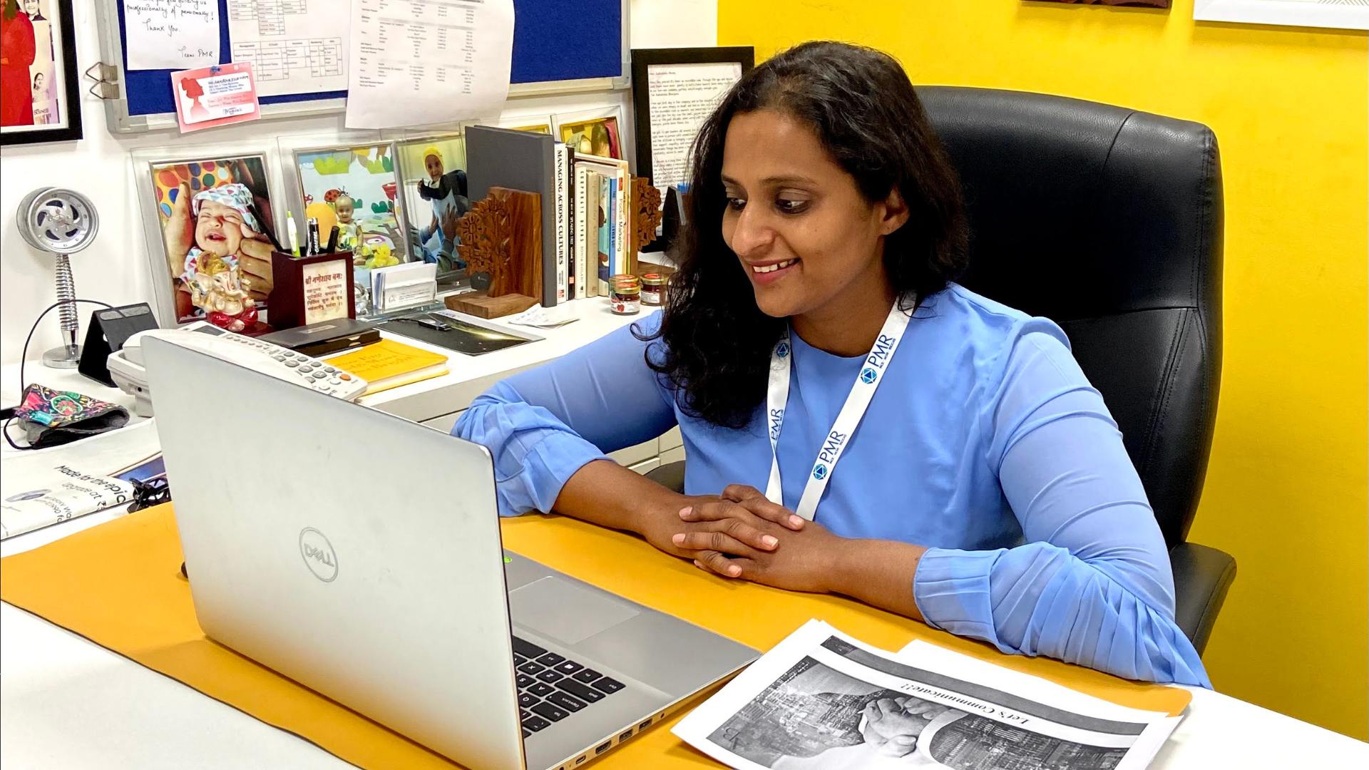 A smiling female entrepreneur wearing a PMR lanyard engages in a video call from her office desk, surrounded by family photos, books, and a Ganesha idol, symbolizing a balance between personal passion, professional commitment, and a love for her work—capturing the spirit of entrepreneurial rebellion.
