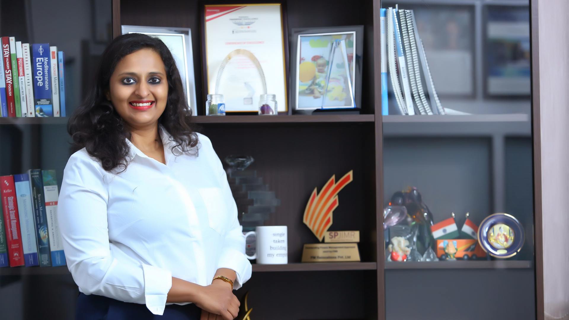 A confident woman entrepreneur in a white shirt stands smiling in front of a bookshelf filled with awards, framed photos, books, and trophies—symbolizing achievement, learning, and the importance of embracing feedback in the journey of business growth.
