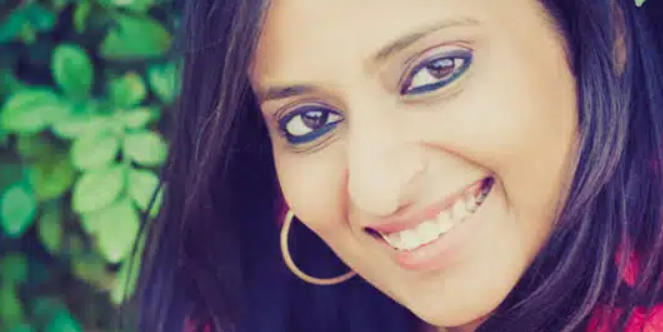 Smiling woman with long black hair and hoop earrings, standing near green foliage, wearing a red top.