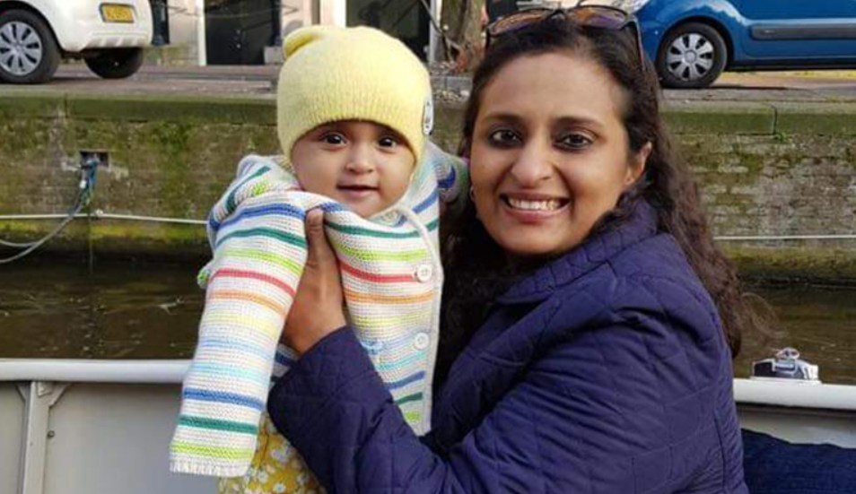 Aakanksha Bhargava smiling and holding her 10-month-old daughter Samaira, who is bundled up in a colorful striped jacket and yellow beanie, during one of their international travels. The background shows a canal and parked cars, suggesting a scenic city location.