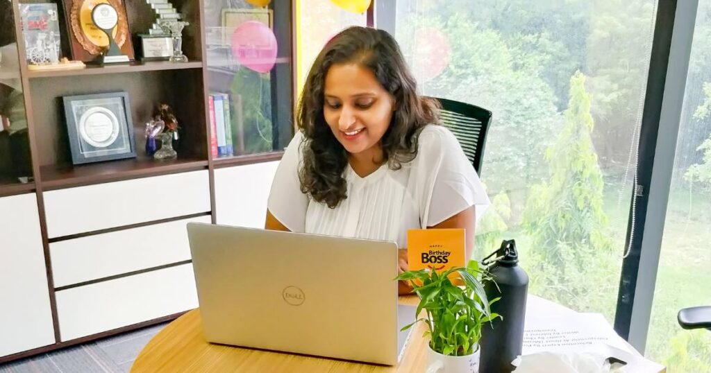 A woman smiling while working on her laptop in a sunlit office with a green view, surrounded by awards, books, a water bottle, a birthday card, and a potted plant on her desk.