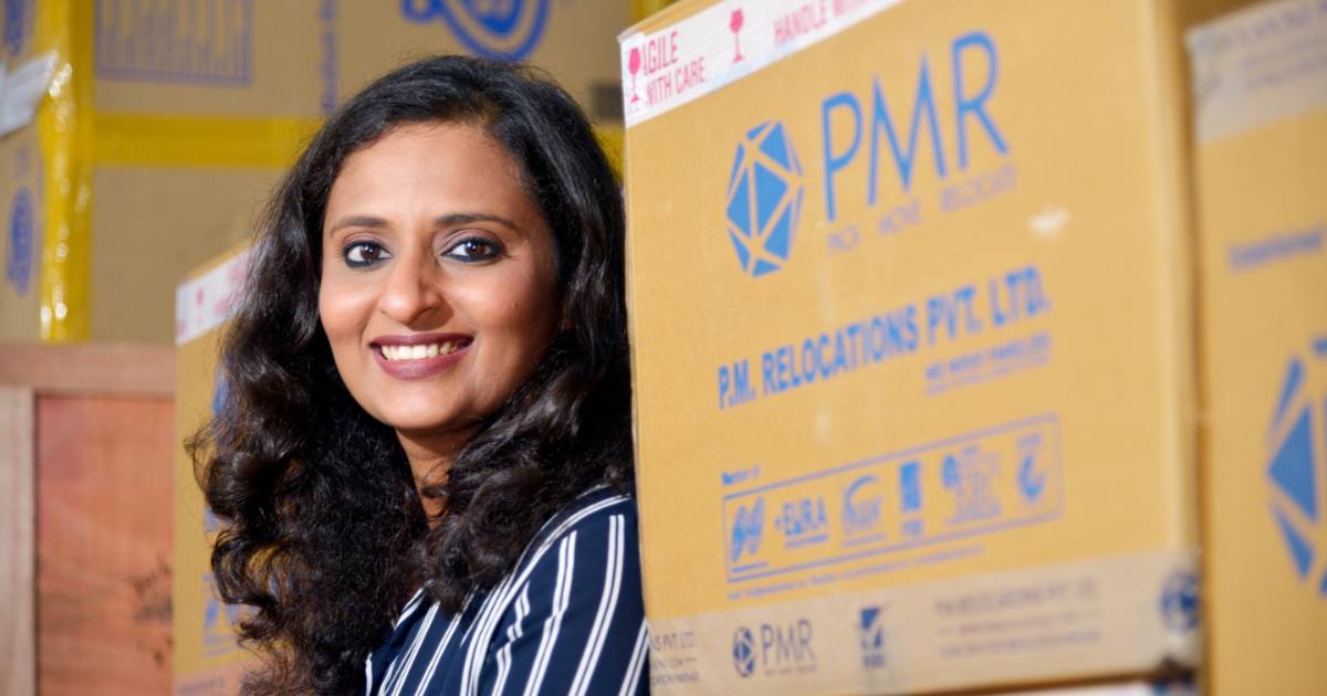 A confident woman with long curly hair smiling beside PMR Relocations Pvt. Ltd. packaging boxes, symbolizing leadership and entrepreneurial success.