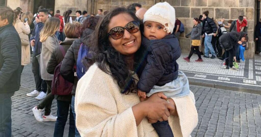 Smiling woman holding a baby in a winter outfit on a busy European cobblestone street, surrounded by tourists and historic architecture.